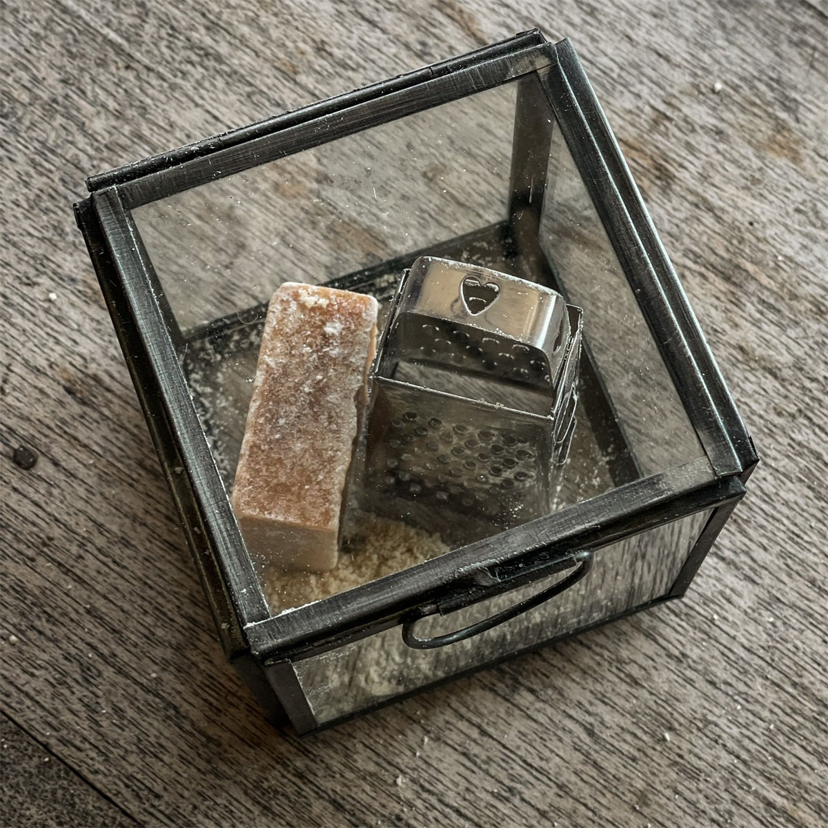 Eine Aufnahme von oben von einem Glaskasten mit einer Raspel in der Farbe silber und einem Duftblock in der Farbe braun